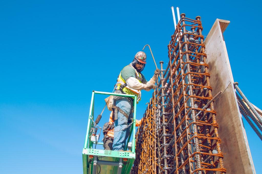 A construction worker in an orange hard hat stands in a green lift while working on a rebar column.